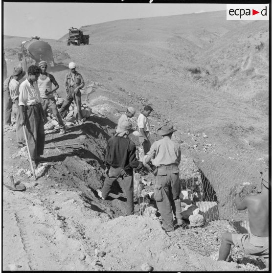 Des ouvriers algériens et des soldats du 9e BG posent des grillage sur le bas-côté de la piste en construction.