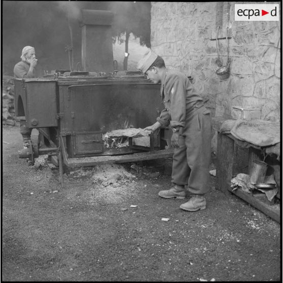 Un tirailleur du 5e bataillon de tirailleurs algériens (BTA) brulant du bois dans une cuisine roulante à Aïn Terzine.