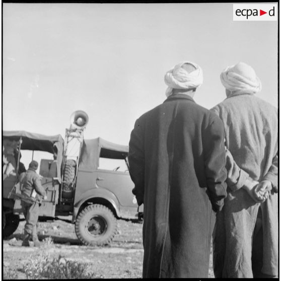 Deux Algériens (de dos) observent le véhicule militaire d'une compagnie de haut-parleurs et de tracts (CHPT) dans un village en Algérie.