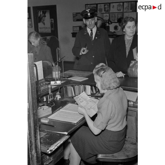 Dans une poste, à un guichet, une femme travaille sous l'oeil d'un homme en uniforme de la Deutsche Reichspost.