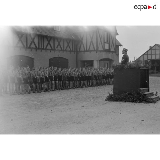 Discours d'un Gruppenführer du N.S.F.K. derrière une tribune ornée du drapeau nazi, en arrière-plan, des dignitaires du N.S.F.K.