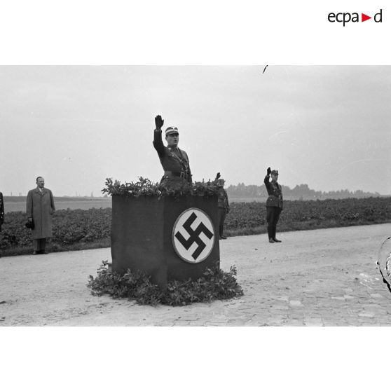 Le Gruppenführer Dr. Otto Zimmermann du N.S.F.K., derrière une tribune, finit un discours et fait le salut nazi.
