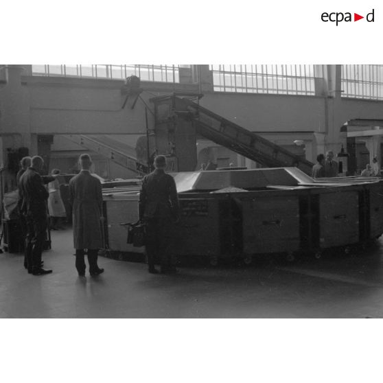 Des ouvriers devant une machine-outil véhiculant des wagonnets à roulettes.