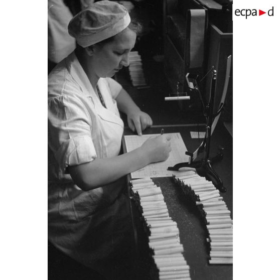 Une femme en blouse blanche, comptabilise et pèse des cigarettes.
