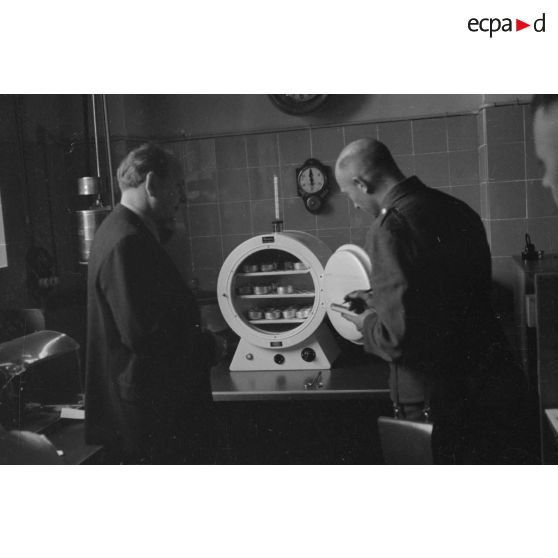 Deux hommes contrôlent le fonctionnement d'une machine (stérilisateur ?) et des objets qu'elle contient.