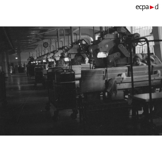 Dans une usine, une rangée de machines-outils actionnées par des femmes.