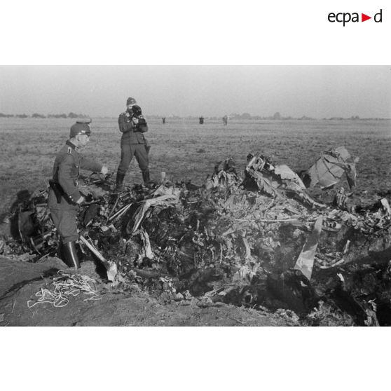 Un policier allemand inspecte des débris d'un avion soviétique abattu, derrière lui un caméraman illustre le sujet pour la propagande.