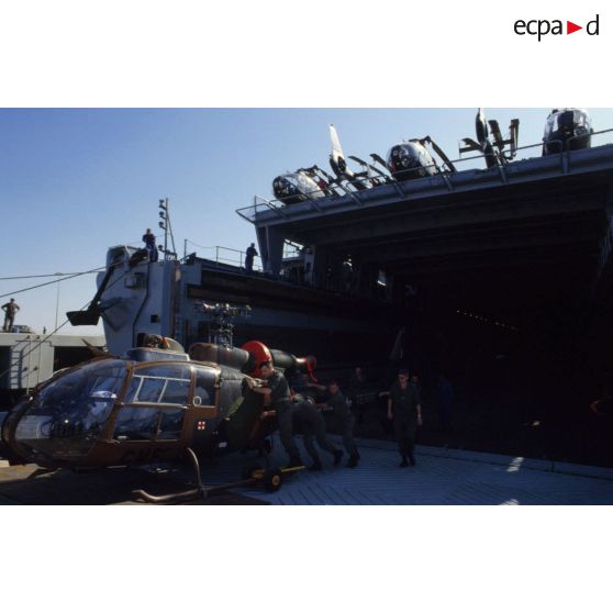 Par la rampe en sortie du hangar du TCD (transport de chalands de débarquement) Foudre, les mécaniciens de l'ALAT (aviation légère de l'armée de terre) déchargent un hélicoptère de combat Gazelle.