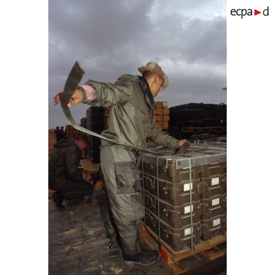 Un soldat portant la tenue S-3P (survêtement de protection à port permanent) (sans l'ANP, appareil normal de protection) tend les sangles de fixation de palettes de munitions (containers de munitions de 5,56 mm pour FAMAS).