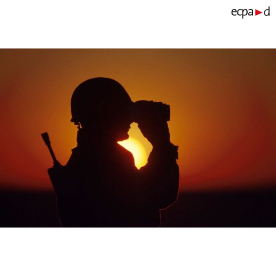 En ZDO (zone de déploiement opérationnel) Olive, un soldat est posté en observation dans le désert, équipé de jumelles, en contre-jour au coucher de soleil.