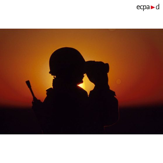 En ZDO (zone de déploiement opérationnel) Olive, un soldat est posté en observation dans le désert, équipé de jumelles, en contre-jour au coucher de soleil.