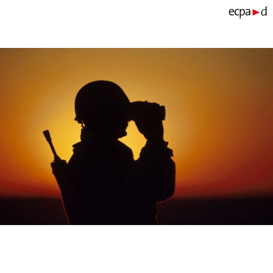 En ZDO (zone de déploiement opérationnel) Olive, un soldat est posté en observation dans le désert, équipé de jumelles, en contre-jour au coucher de soleil.