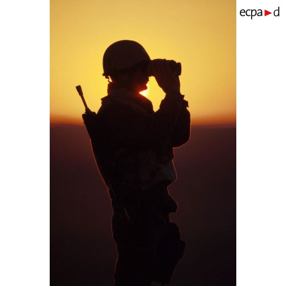 En ZDO (zone de déploiement opérationnel) Olive, un soldat est posté en observation dans le désert, équipé de jumelles, en contre-jour au coucher de soleil.