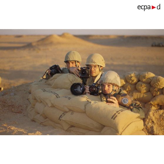 En ZDO (zone de déploiement opérationnel) Olive, trois reporters photographes de l'ECPA (Etablissement cinématographique et photographique des Armées) posent postés derrière un abri de sacs de sable.
