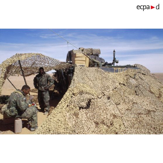 En ZDO (zone de déploiement opérationnel) Olive, un groupe de soldats américains prend son repas au pied d'un transport de troupes M-113, sous un filet de camouflage.