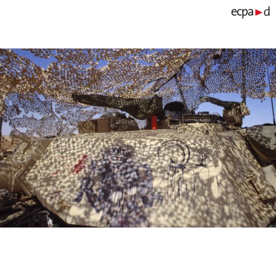 En ZDO (zone de déploiement opérationnel) Olive, tourelle d'un char de combat Abrams-M1 américain, décorée d'un dessin représentant la mort tenant une hache et un loup sur une falaise au clair de lune, sous un filet de camouflage.