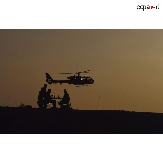 En ZDO (zone de déploiement opérationnel) Olive, deux joueurs d'échecs du 6e REG (régiment étranger du génie) assis à une table, survolés par un hélicoptère de combat Gazelle, en contre-jour dans le soleil couchant.