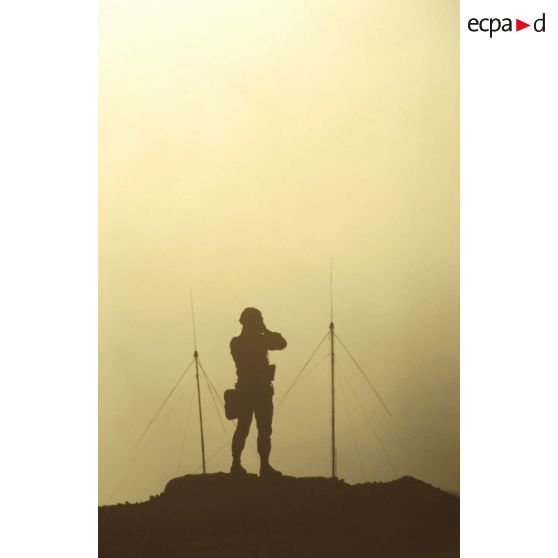 Au soleil couchant, un soldat, se détachant en ombre chinoise, observe aux jumelles.