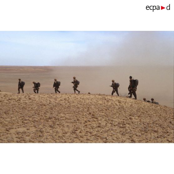En ZDO (zone de déploiement opérationnel) Olive, des soldats marchent en colonne dans le désert, en tenue de combat.