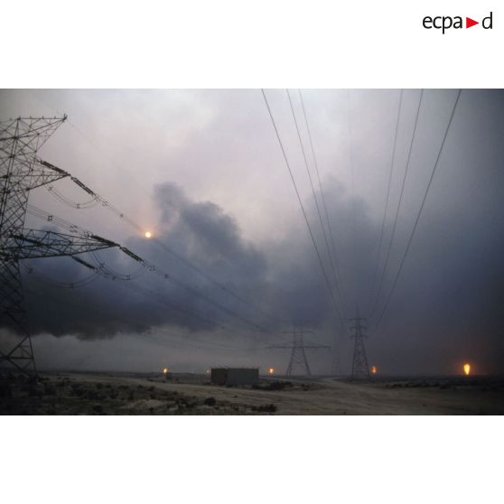 Puits de pétrole en feu derrière des pylones électriques.
