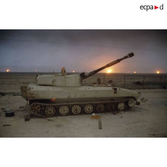 Un canon automoteur irakien de 122 mm M1974 (ou 2S1) abandonné devant un horizon de puits de pétrole en feu.