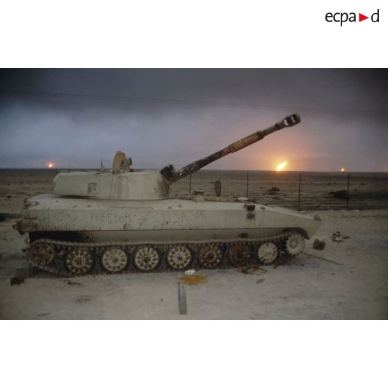 Un canon automoteur irakien de 122 mm M1974 (ou 2S1) abandonné devant un horizon de puits de pétrole en feu.