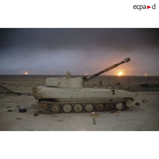 Un canon automoteur irakien de 122 mm M1974 (ou 2S1) abandonné devant un horizon de puits de pétrole en feu.
