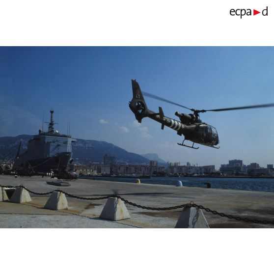 Un hélicoptère de combat Gazelle du 3e RHC (régiment d'hélicoptères de combat) décolle d'un quai du port de Toulon. En arrière-plan le TCD (transport de chalands de débarquement) Foudre est amarré au quai.