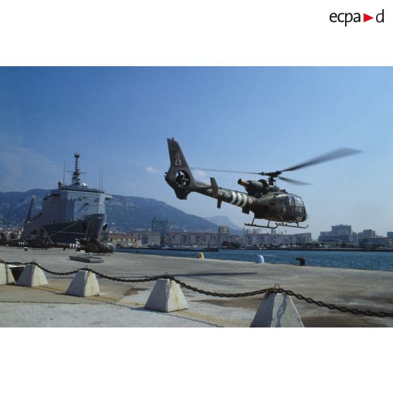 Un hélicoptère de combat Gazelle du 3e RHC (régiment d'hélicoptères de combat) décolle d'un quai du port de Toulon. En arrière-plan le TCD (transport de chalands de débarquement) Foudre est amarré au quai.