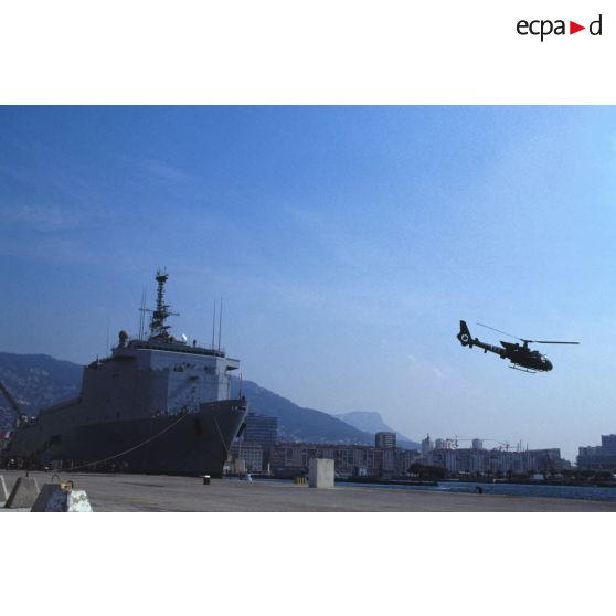 Un hélicoptère de combat Gazelle du 3e RHC (régiment d'hélicoptères de combat) décolle d'un quai du port de Toulon. En arrière-plan le TCD (transport de chalands de débarquement) Foudre est amarré au quai.