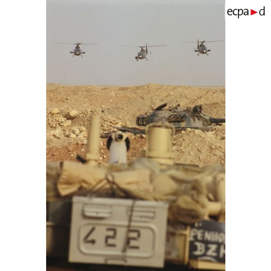 Des hélicoptères de combat Gazelle Hot et Canon volent vers l'objectif avec au premier plan un blindé de reconnaissance AMX-10 RC. Au second plan une tourelle de char de combat T-55 irakien.