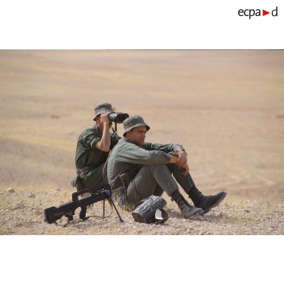 Soldats scrutant le désert à l'aide de jumelles, assis à côté d'un fusil d'assaut FAMAS.