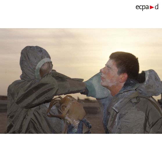 Un soldat en tenue S-3P (survêtement de protection à port permanent) et portant l'ANP (appareil normal de protection) nettoie le visage de son camarade à l'aide d'un gant de décontamination chimique d'urgence lors de l'exercice NBC du 1er RI.
