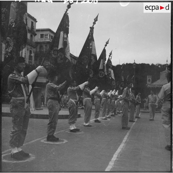 Porte-drapeaux devant le monument aux morts et deux soldats présentant chacun un coussin de décorations.
