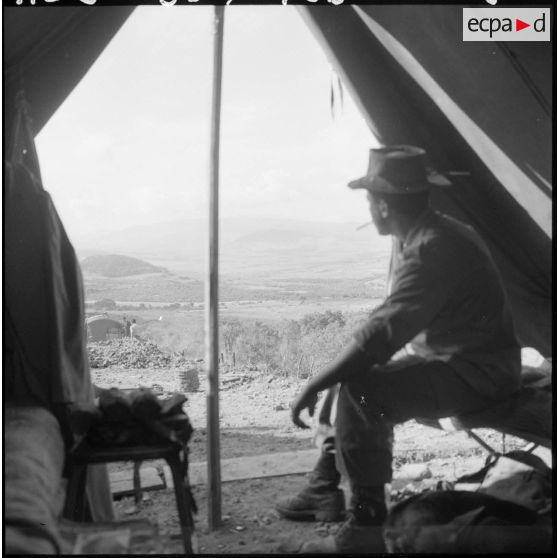 Portrait d'un militaire qui fume une cigarette en contemplant la vie quotidienne du camp, assis à l'intérieur de sa tente.