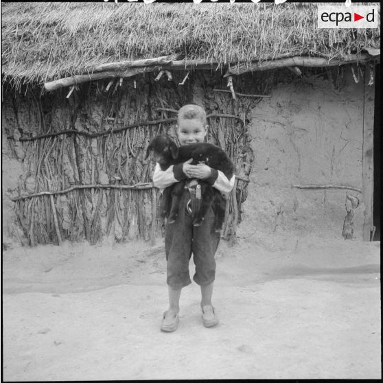 Un petit garçon devant sa mechta portant dans ses bras un petit agneau.