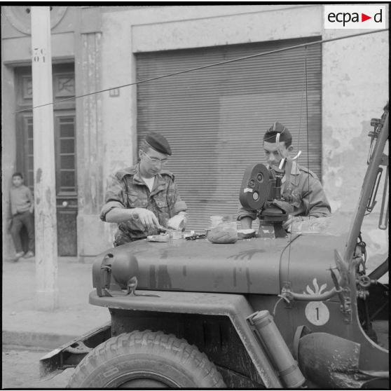 Alger. Belcourt. Un soldat et un caméraman du Service cinématographique des armées (SCA) mangent sur le capot d'une jeep pendant les manifestations.