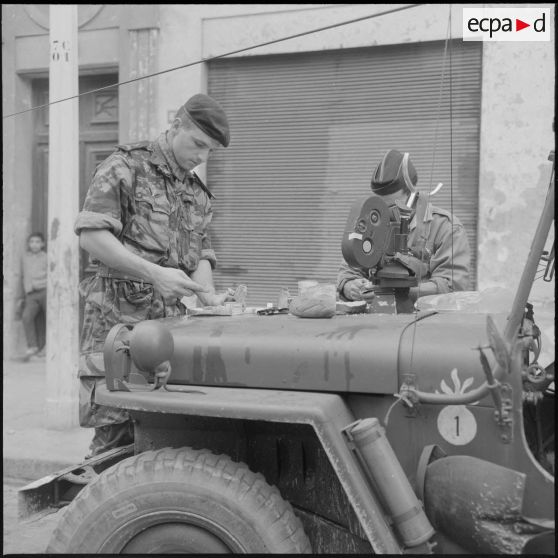 Alger. Belcourt. Un soldat et Alain Darchy, caméraman du Service cinématographique des armées (SCA), mangent sur le capot d'une jeep pendant les manifestations.