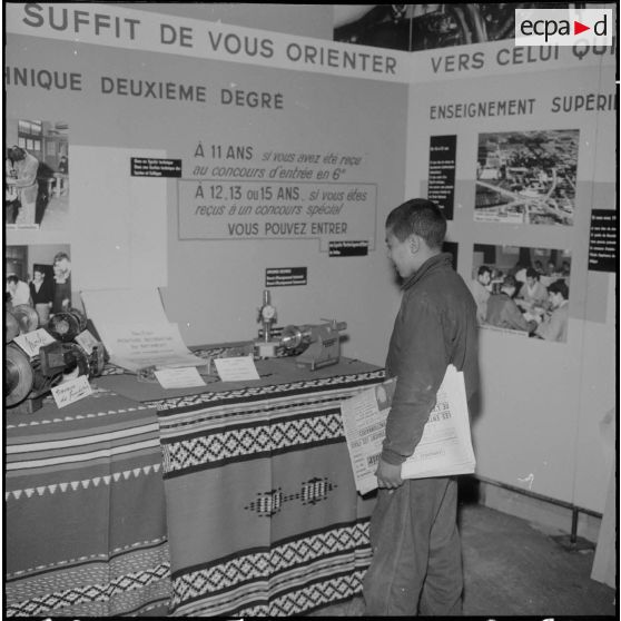 Inauguration de la foire exposition d'Oran. Un jeune vendeur de journaux devant une exposition de réalisations de jeunes apprentis, au pavillon de l'orientation professionnelle.
