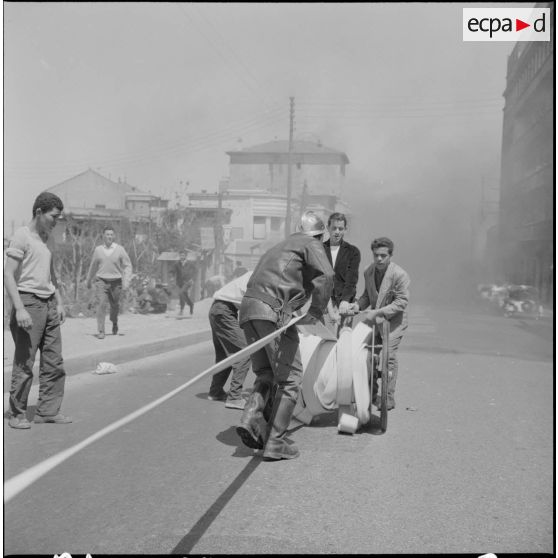 Alger. Quartier des Tagarins. Explosion d'un camion-citerne. Des civils aident un pompier à dérouler la lance à incendie.