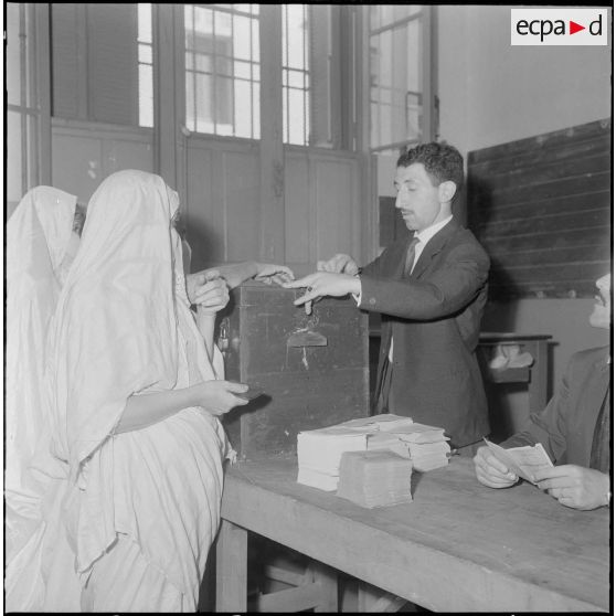 Journée de vote pour le référendum d'autodétermination (1er juillet 1962). Une femme glisse son bulletin de vote dans l'urne face à un assesseur du bureau de vote n°1.