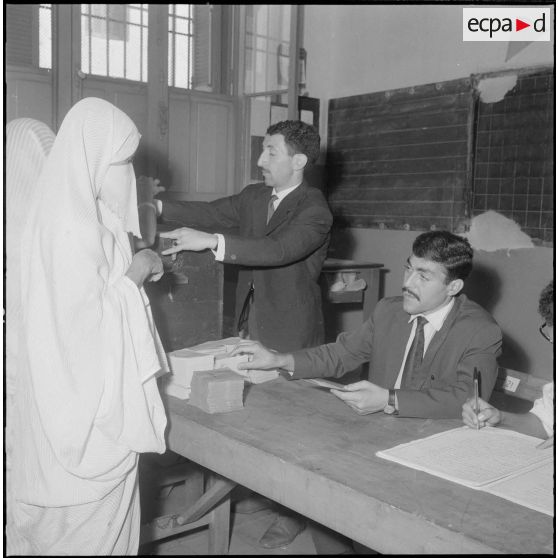 Journée de vote pour le référendum d'autodétermination (1er juillet 1962). Une femme glisse son bulletin de vote dans l'urne face à un assesseur du bureau de vote n°1.
