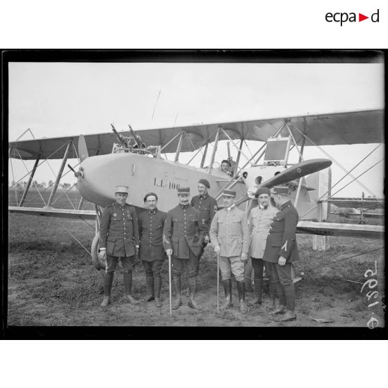[Des autorités militaires posent devant un avion militaire équipé de mitrailleuses, sur un camp d'aviation.]