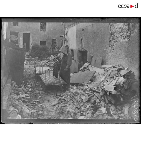 [Vue d'une femme au milieu de débris, devant une maison où sont disposés deux lits ; un petit balais en paille à la main.]