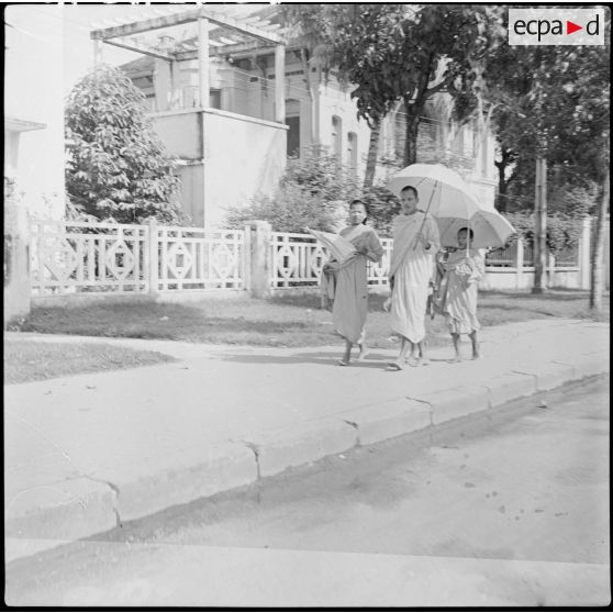 1 Trois jeunes bonzes, portant une ombrelle, marchent pieds nus les uns derrière les autres dans une rue. [description provisoire]