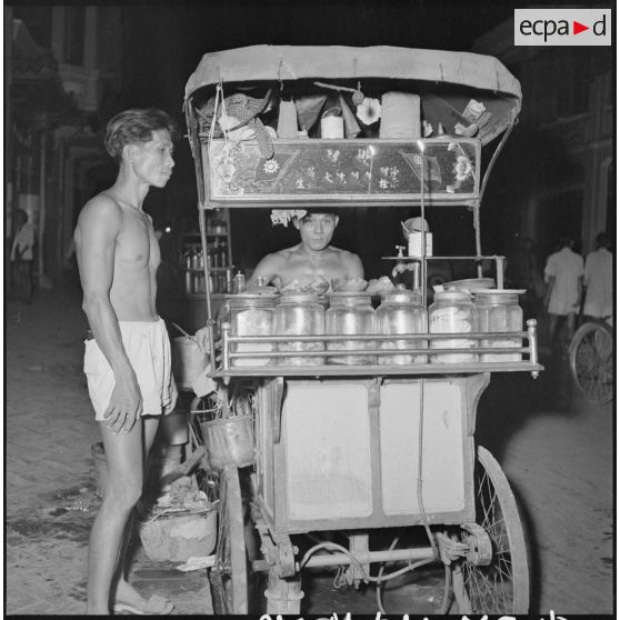 Dans une rue de Phnom Penh, un commerçant ambulant derrière sa charrette sur laquelle il transporte et présente ses marchandises. [description provisoire]