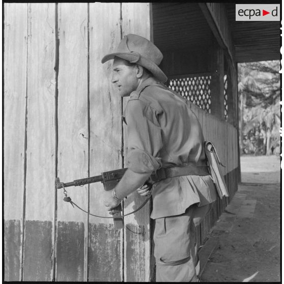 26 Le sergent Julien Mosca avec un PM MAT 49, posté derrière un mur, surveille la fouille d'un camp viêt minh abandonné. [description provisoire]