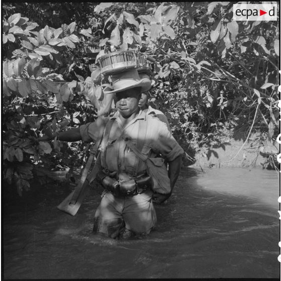 Franchissement d'un stung par un soldat du 2/6e RIC (2e bataillon du 6e Régiment d'Infanterie Coloniale). Ce tirailleur porte sur sa tête, posés en équilibre, deux boîtiers chargeurs de 150 cartouches pour mitrailleuse modèle 1931 ""Reibel"" de calibre 7,5 mm. [description provisoire]