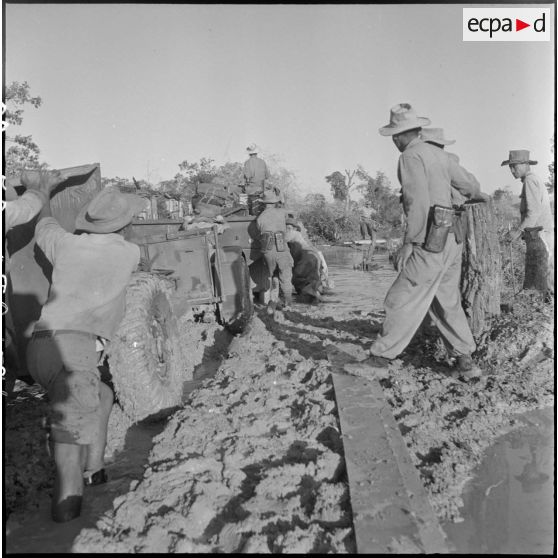Un convoi de GMC 6x6 (General Motors Corporation) est ralenti par les coupures de la piste et les inondations. Plusieurs ponts ont été brûlés par le Viêt-minh. Quelques jours auparavant, des dérivations avaient été aménagées mais suite à de récents orages, les abords ont été inondés et les rampes transformées en torrents boueux. Ici, la pièce d'artillerie a peine à se dégager de la boue. [description provisoire]