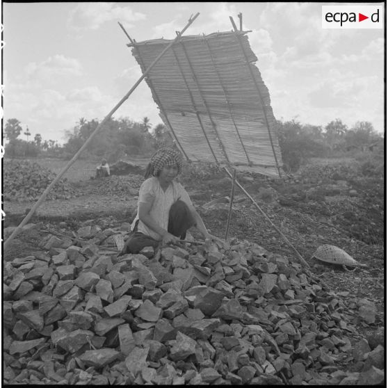 Abritée derrière un pare-soleil, une femme cambodgienne casse des pierres avec un petit marteau, pour le terrassement de la route. [description provisoire]